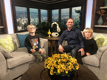 One adult and two children sit on couches in a television studio with Portland in the background.