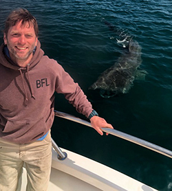 Man with sandy hair stands on the left side of the frame wearing a brown sweatshirt with BFL on the chest. A large shark is underwater in the right side of the photo.