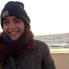 Smiling person with dark hair wears a Carhartt beanie on the beach.